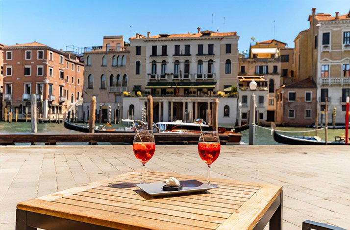 Two Aperol Spritz cocktails with cicchetti on an outdoor table overlooking Venice's Grand Canal near Rialto.