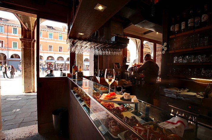 Bartender preparing drinks behind a cicchetti counter at Osteria Bancogiro, with wine glasses overhead and a Venice piazza visible beyond.