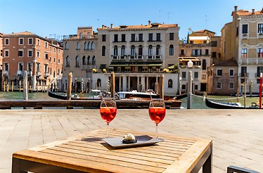 Two Aperol Spritz cocktails with cicchetti on an outdoor table overlooking Venice's Grand Canal near Rialto.