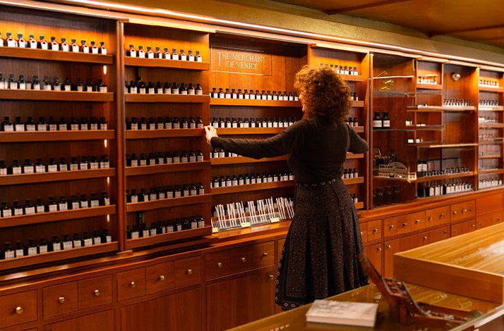 Woman reaching into the perfumer's organ at The Merchant of Venice's Sala Profumo workshop, walls lined with labeled bottles.