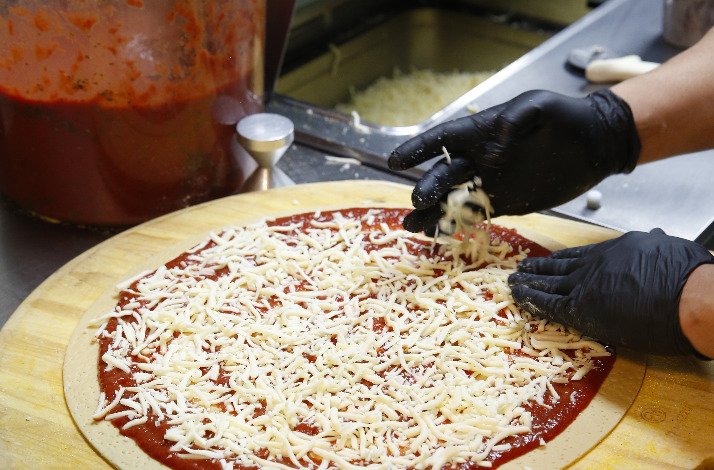 Gloved hands spreading shredded mozzarella over tomato-sauced pizza dough on a wooden peel at Novel Pizza Cafe.