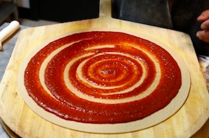 Tomato sauce spread in a spiral on thin pizza dough resting on a wooden peel, ready for topping at Novel Pizza Cafe.
