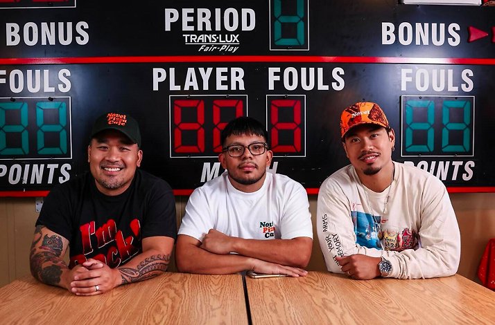 Three Novel Pizza Cafe team members seated at a table, posing in front of a vintage basketball scoreboard wall.