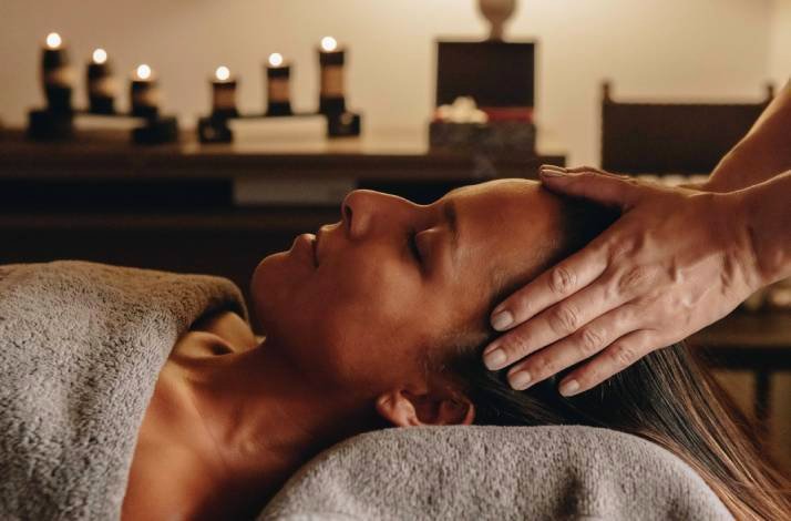 Woman reclining with eyes closed, receiving a soothing head massage in a warmly candlelit spa treatment room.