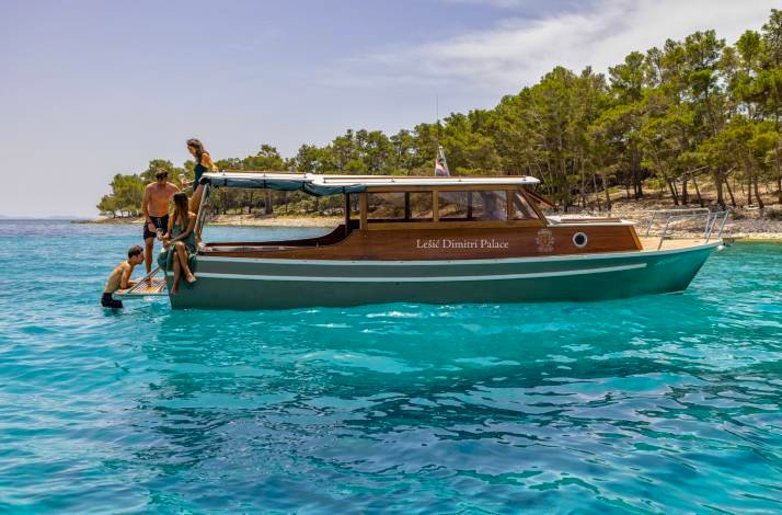 Guests boarding the Kata, a restored wooden boat marked 'Lešić Dimitri Palace,' anchored in crystal-clear turquoise Adriatic waters.