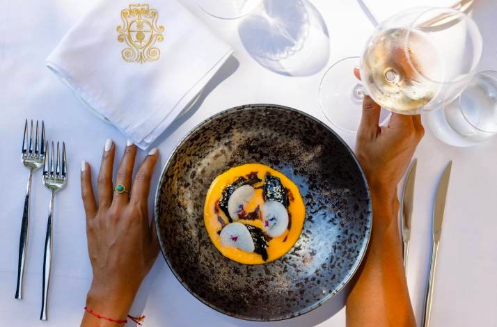 Overhead view of a plated tasting-menu course at LD Restaurant, with a wine glass held alongside and a monogrammed napkin nearby.
