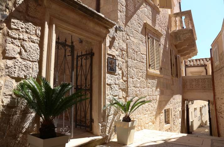 Sun-drenched stone alleyway at Lešić Dimitri Palace in Korčula Old Town, framed by ornate iron gates and potted palms.