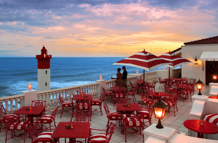Oceanfront terrace dining with red tables, striped umbrellas, and a lighthouse at sunset.