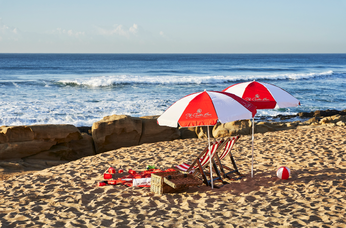 Beach picnic setup with two red-and-white umbrellas beside rocky shore and waves.
