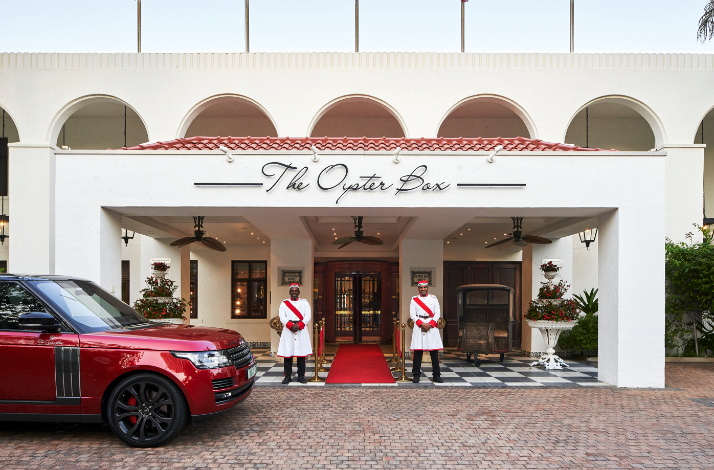 The Oyster Box hotel entrance with doormen on a red carpet beneath arched portico.