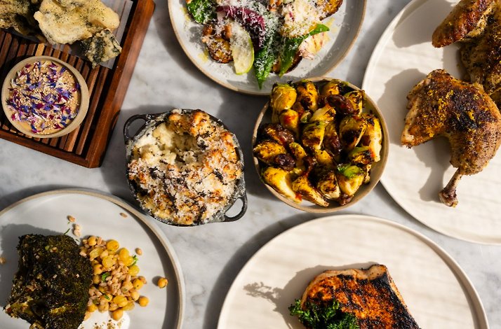 Farm-to-table feast at North Fork Table & Inn showcasing roasted chicken, charred vegetables, baked mac and cheese, and herb-crusted fish on marble surface.