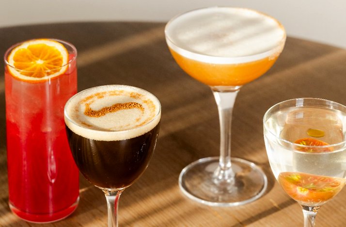 Assortment of handcrafted cocktails at North Fork Table & Inn, garnished with orange slice, edible flowers, and spiced foam, resting on a wood table.