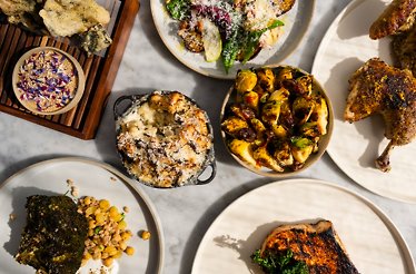 Farm-to-table feast at North Fork Table & Inn showcasing roasted chicken, charred vegetables, baked mac and cheese, and herb-crusted fish on marble surface.