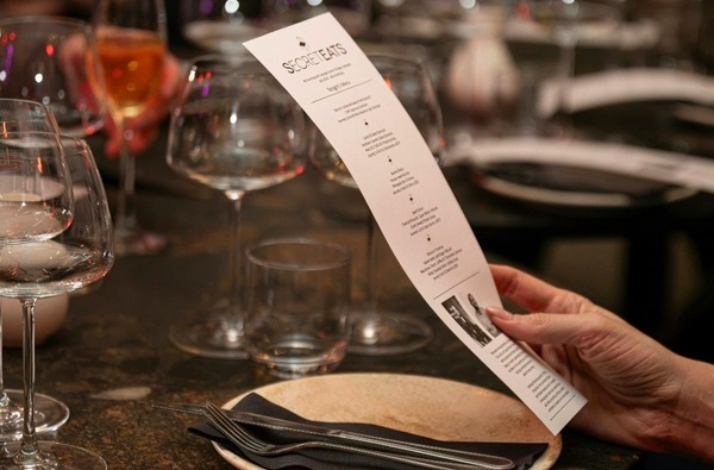 Guest holding a printed SecretEATS menu at a candlelit table with wine glasses.
