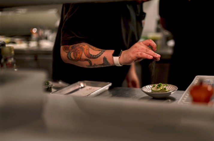 Chef with octopus forearm tattoo finishing a plated dish with a pinch of seasoning at Zitz Sum.