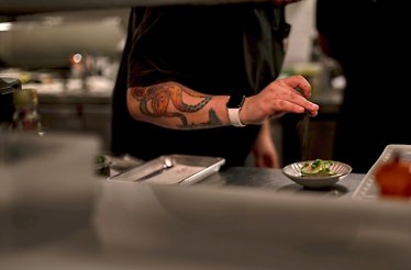 Chef with octopus forearm tattoo finishing a plated dish with a pinch of seasoning at Zitz Sum.