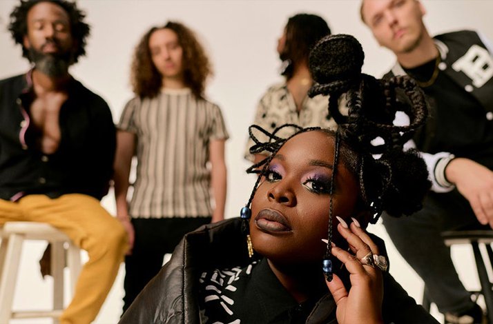 Tank of Tank and the Bangas posed in the foreground, with four band members standing in soft-lit studio background.