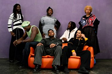 Members of KOKOROKO posing together on and around an orange sofa against a purple wall, wearing eclectic outfits.