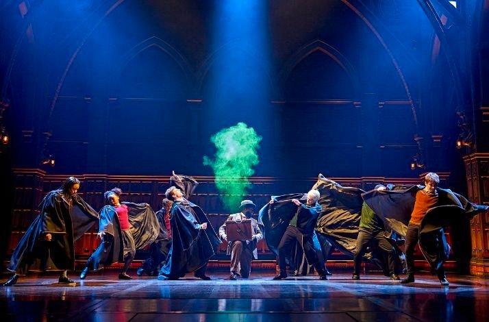 Ensemble cast in dark robes performs a spell as green smoke rises center stage under dramatic blue lighting at a Harry Potter and the Cursed Child showing.