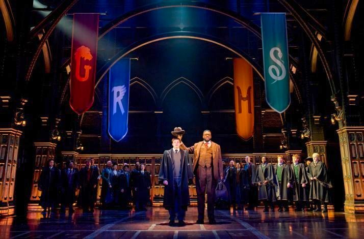 Two robed students stand front and center in a Hogwarts hall with house banners overhead and a crowd of witches and wizards behind them at a Harry Potter and the Cursed Child showing.