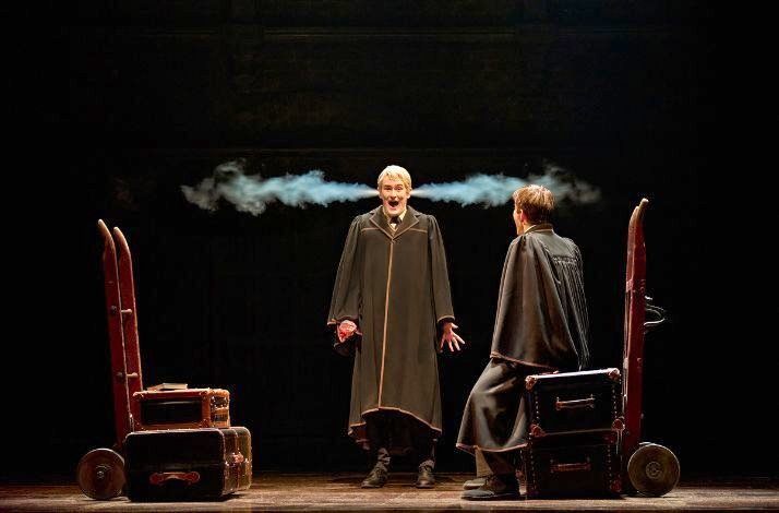 Two cloaked men face each other between wheeled trunks as a burst of smoke shoots from one man’s ears in a dark, spotlighted scene at a Harry Potter and the Cursed Child showing.