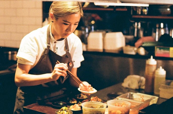 Chef Esther Choi plating dishes with chopsticks in a restaurant kitchen, wearing a brown apron over a white chef's coat.