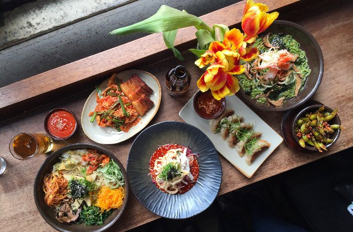 Overhead spread of Korean dishes by chef Esther Choi, including bibimbap, dumplings, pork belly, and noodles, on a wooden table. 