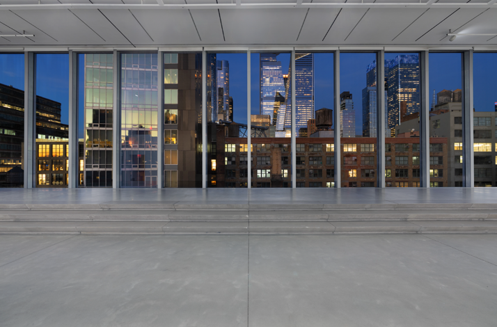 Spacious empty gallery interior with track lighting, white walls, and a stepped platform before floor-to-ceiling windows with NYC skyline at dusk.