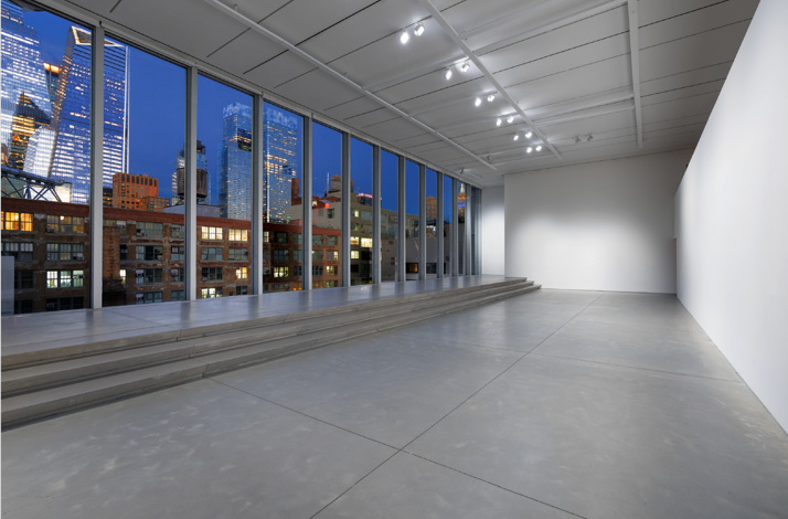 Empty gallery space with stepped concrete platform facing floor-to-ceiling windows overlooking an illuminated NYC skyline at dusk.