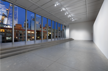 Empty gallery space with stepped concrete platform facing floor-to-ceiling windows overlooking an illuminated NYC skyline at dusk.