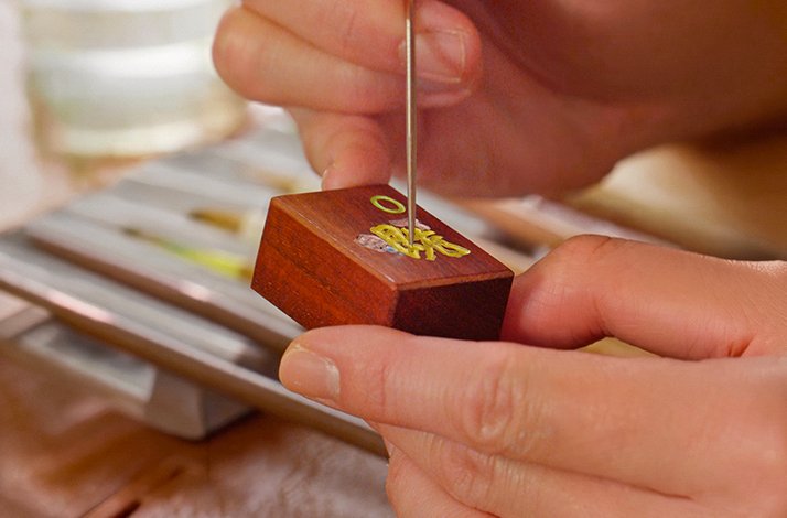 Artisan hand-engraving a wooden mahjong tile with gold Chinese characters using a fine tool.