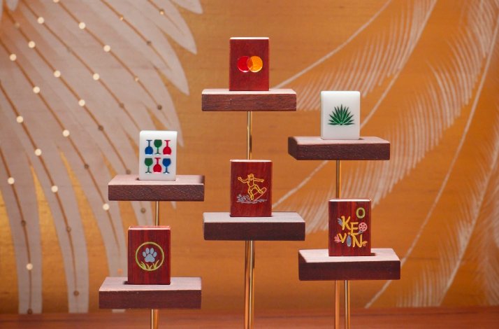 Six personalised handcrafted mahjong tiles on wooden stands, featuring a Mastercard logo, paw print, name 'Kevin', and custom motifs.