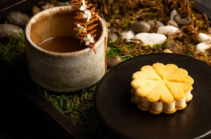 Flower-shaped golden waffle sandwich cookie on a black plate beside a rustic ceramic broth cup, set on a moss-and-stone tray at Nightbird.