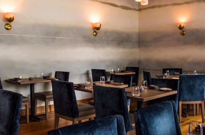 Elegantly set dining room at Nightbird in Hayes Valley, with plush navy seating, natural wood tables, and soft amber sconce lighting on a painted grey wall.