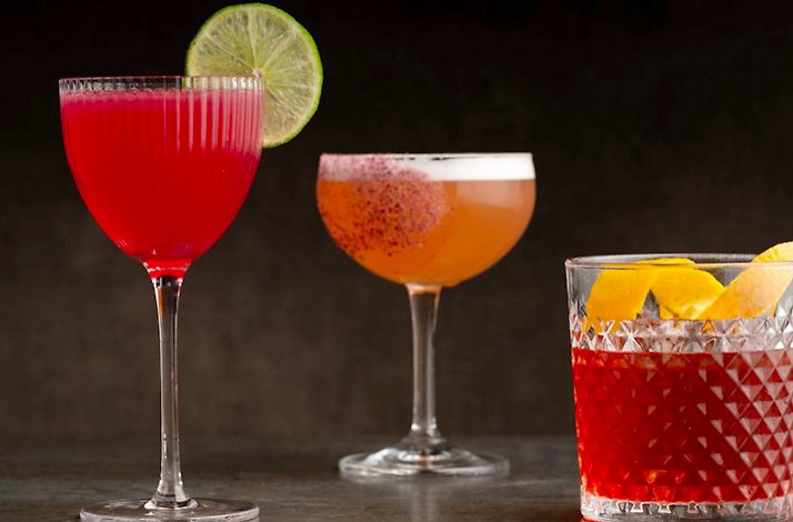 Trio of artisan cocktails at Nightbird, featuring vibrant red, amber, and dark ruby drinks garnished with lime, spiced rim, and orange peel.