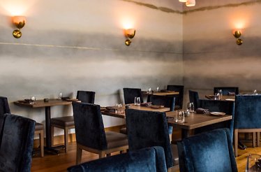 Elegantly set dining room at Nightbird in Hayes Valley, with plush navy seating, natural wood tables, and soft amber sconce lighting on a painted grey wall.