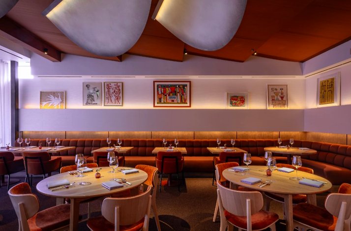 Jacaranda dining room with rust-velvet banquette seating, round oak tables, sculptural ceiling panels, and framed artworks.