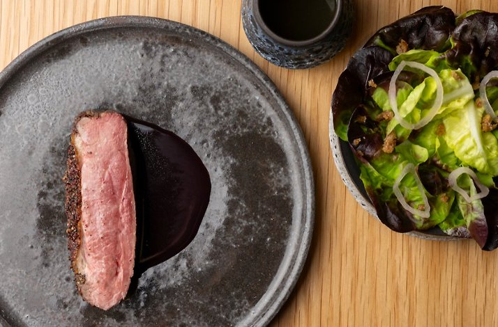 Sliced pepper-crusted meat resting on dark sauce, plated alongside a mixed leaf and shallot salad at Jacaranda.