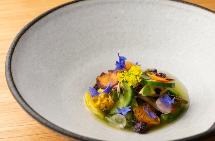 Seasonal vegetables and edible blooms arranged in a light herb broth, served in a dark-rimmed ceramic bowl at Jacaranda.