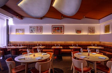 Jacaranda dining room with rust-velvet banquette seating, round oak tables, sculptural ceiling panels, and framed artworks.