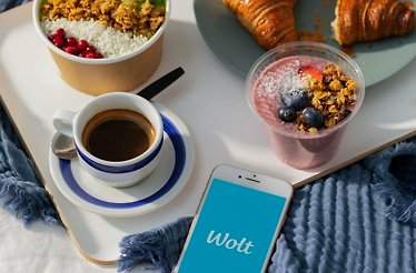 Wolt app displayed on smartphone beside breakfast tray with coffee, acai smoothie bowl, granola bowl, and croissants.