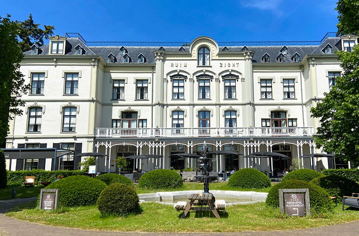 Exterior of Hotel Villa Ruimzicht showing ornate historic facade, landscaped garden with fountain, and outdoor terrace seating.