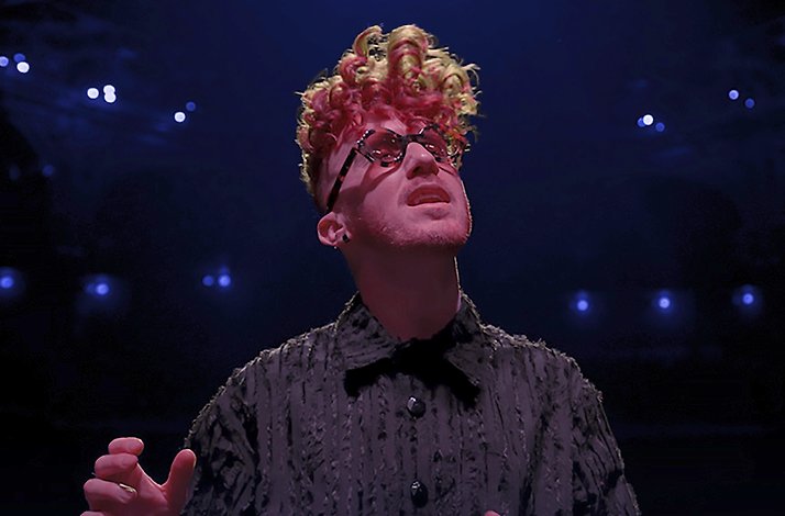 Musician Daley performing on stage with red and blonde curly hair, dark glasses, and a textured black shirt.