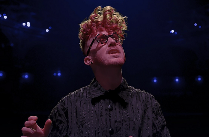 Musician Daley performing on stage with red and blonde curly hair, dark glasses, and a textured black shirt.