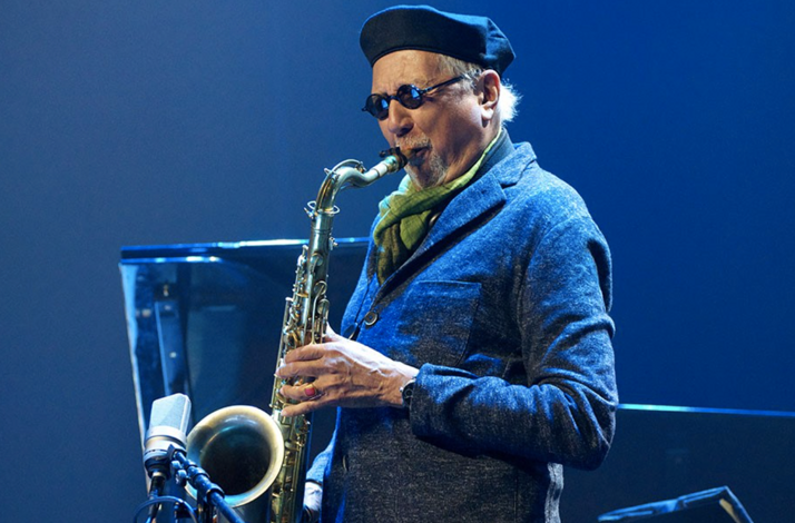 Musician Charles Lloyd playing a saxophone against a solid blue background.