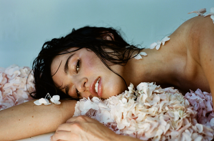 Singer Rozzi lying with wet black hair amidst pale pink flowers against a light blue background.