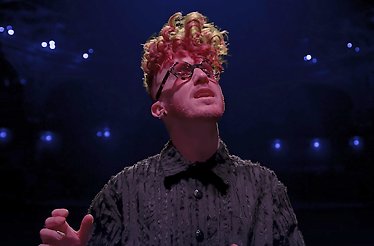 Musician Daley performing on stage with red and blonde curly hair, dark glasses, and a textured black shirt.