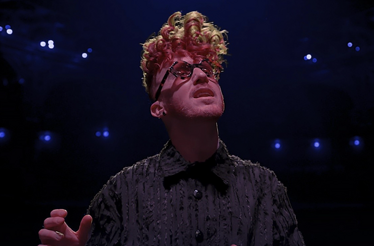 Musician Daley performing on stage with red and blonde curly hair, dark glasses, and a textured black shirt.