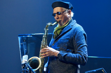 Musician Charles Lloyd playing a saxophone against a solid blue background.
