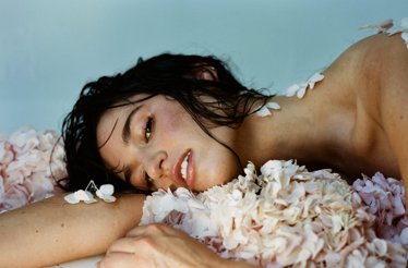 Singer Rozzi lying with wet black hair amidst pale pink flowers against a light blue background.